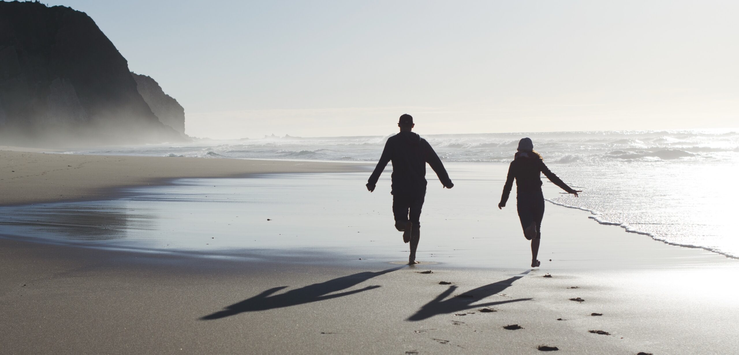 Couple running on a sandy beach with a cliff in the background.
