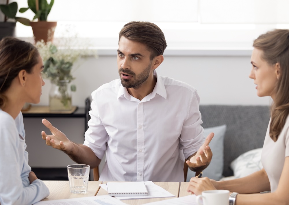Man gesturing emphatically during a tense leadership discussion with two colleagues.