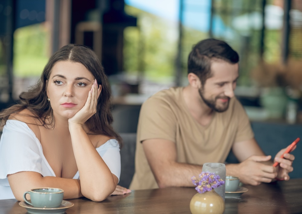 Woman looking bored while man on phone at table, wrong partner choice