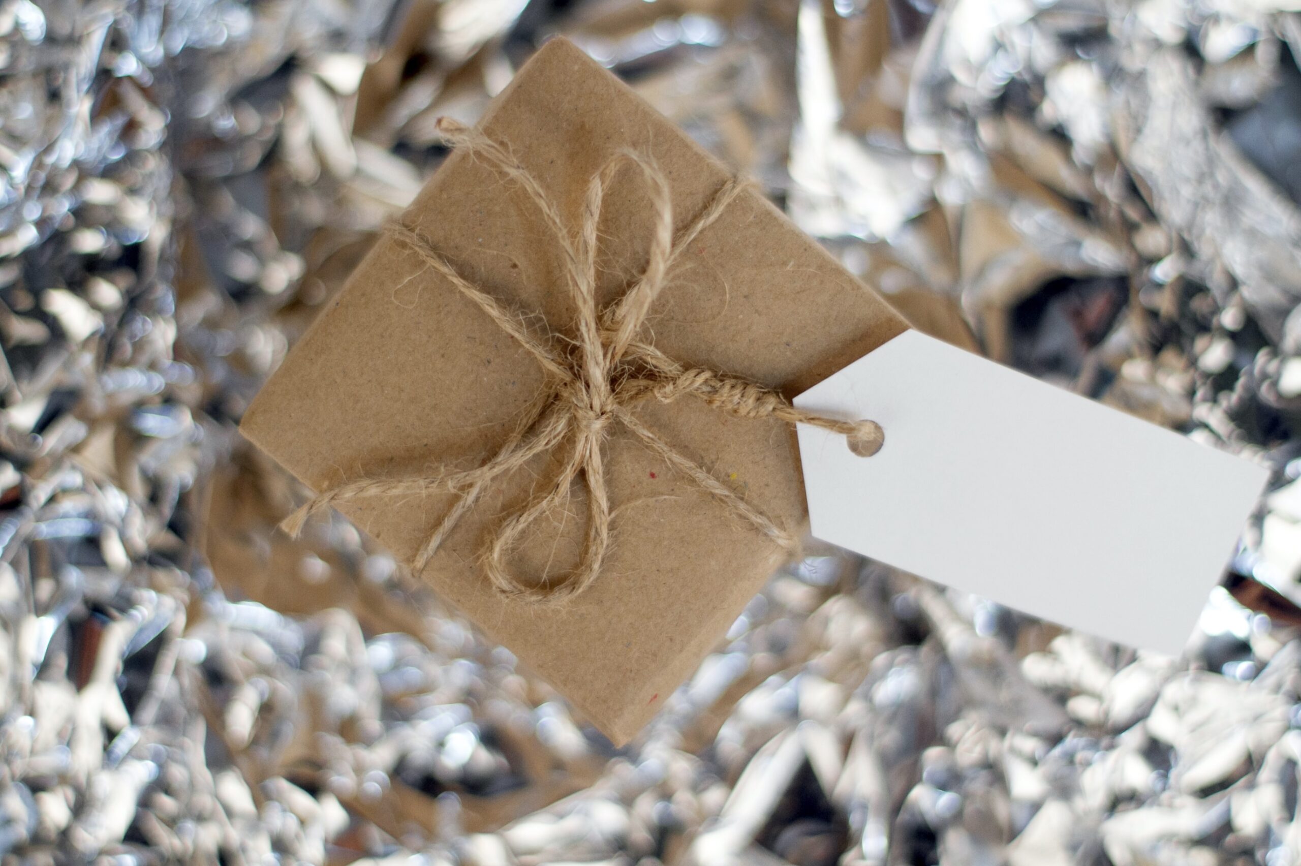 Brown paper gift box with twine bow and blank white tag on shimmering silver background.